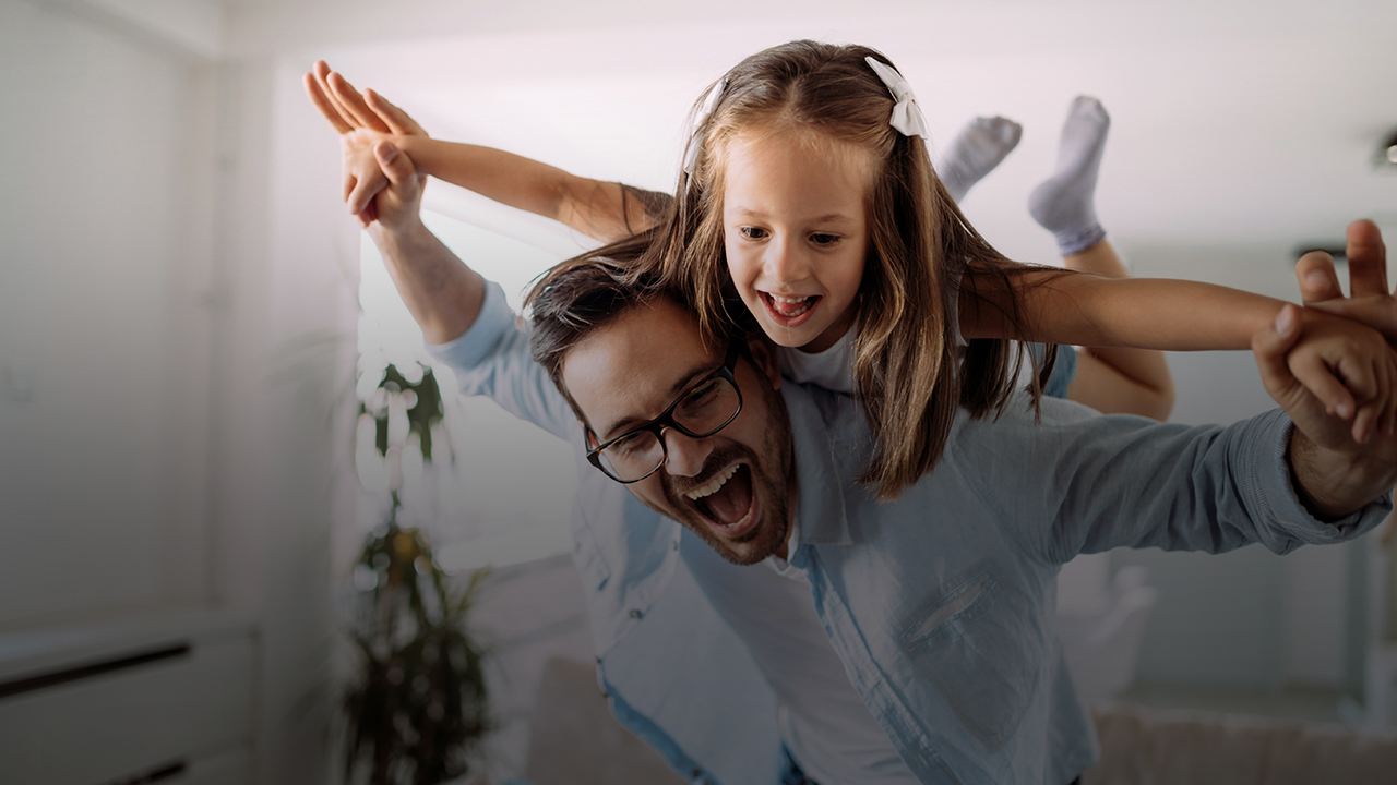 Ein Mädchen liegt mit dem Bauch auf der Schulter ihres Vaters. Sie haben beide die Hände gestreckt und spielen Flugzeug.